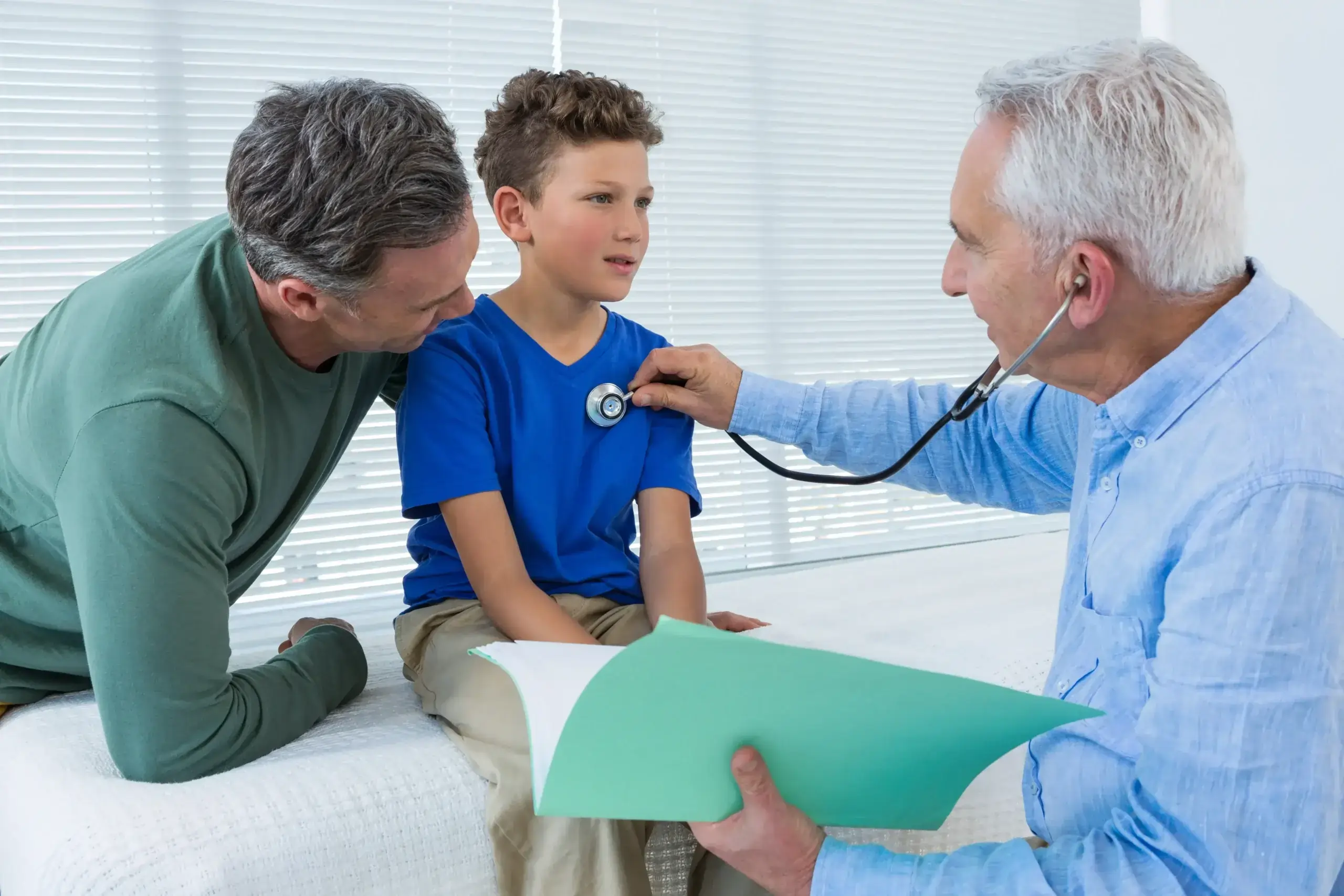 father looking at son while son being examine by doctor with stethoscope on his routine physical checkup