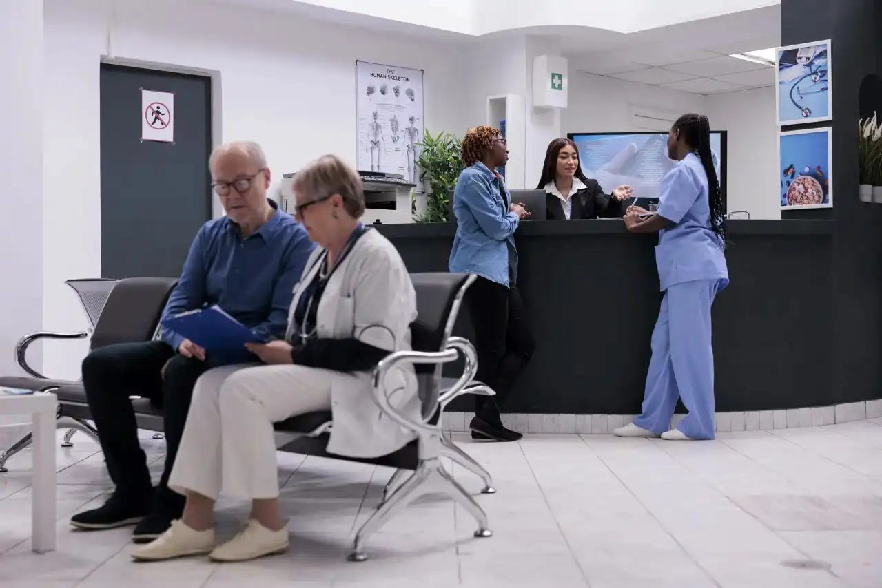 doctor sitting near an old patient discussing his walk-in appointment while a receptionist behind her discussing upcoming appointment with her colleagues